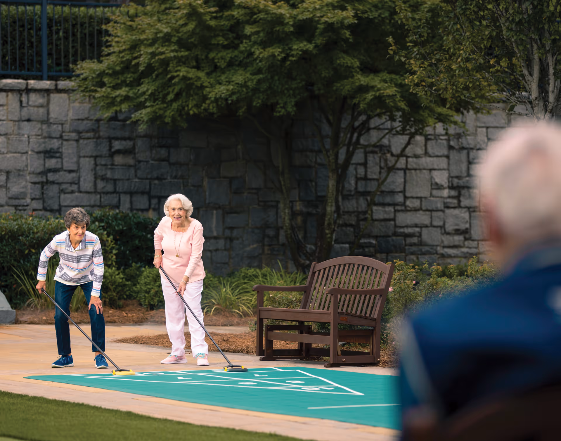Two elderly women playing shuffleboard outdoors on a green shuffleboard court, with a stone wall and trees in the background. A wooden bench is nearby, and a person is partially visible in the foreground watching the game.