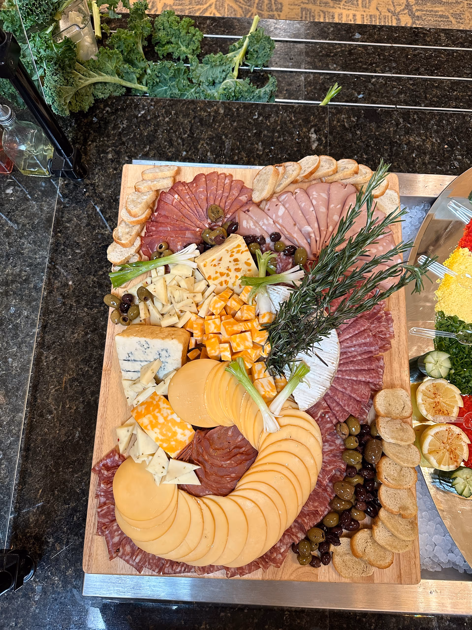 A wooden platter with an assortment of sliced cheeses, cured meats, olives, green onions, and small toasted bread slices arranged decoratively. The platter is placed on a dark granite countertop with some kale leaves in the background and part of another food tray visible on the right side.