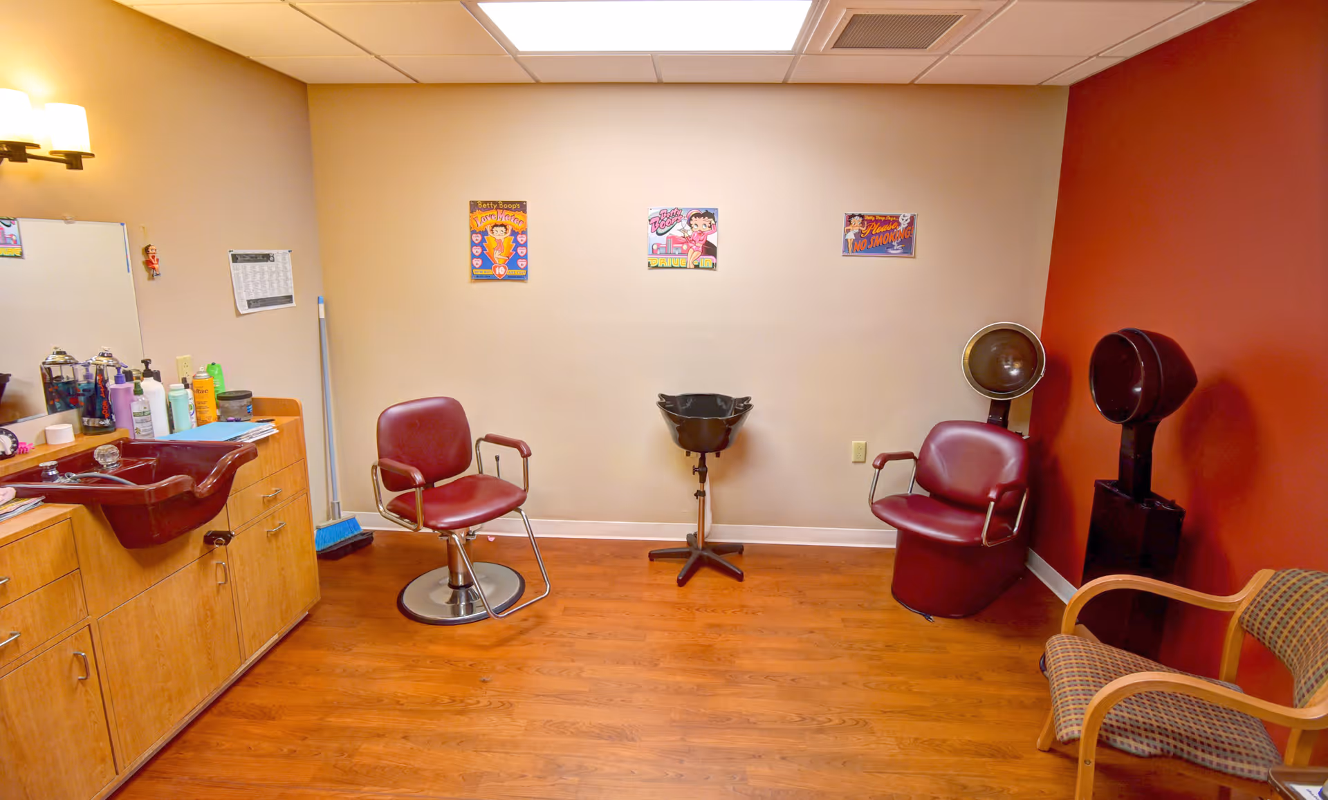 Small salon room with styling chairs, a shampoo sink, hair dryers and cabinets against beige and red walls.
