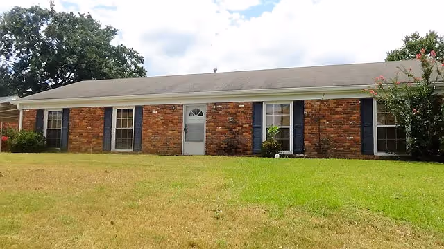 Single-story brick residential building with a gray shingled roof, white door with a window, and multiple windows with dark shutters. The building is surrounded by a grassy lawn with some trees and bushes.