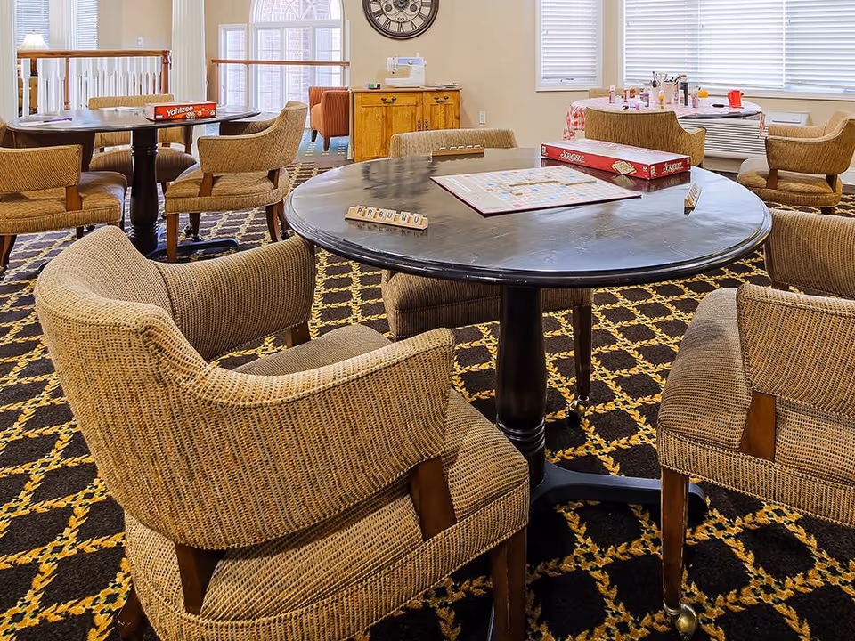 A cozy game room with round tables and wicker chairs arranged on a patterned carpet. Board games including Scrabble and Yahtzee are set up on the tables. The room has large windows with blinds, a wooden cabinet, and a large wall clock.