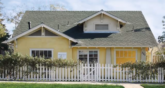 Front exterior view of a single-story yellow house with a green shingled roof, white trim, and a white picket fence in front. There are bushes along the fence and a small sign on the house that reads 'Mission Villa'.
