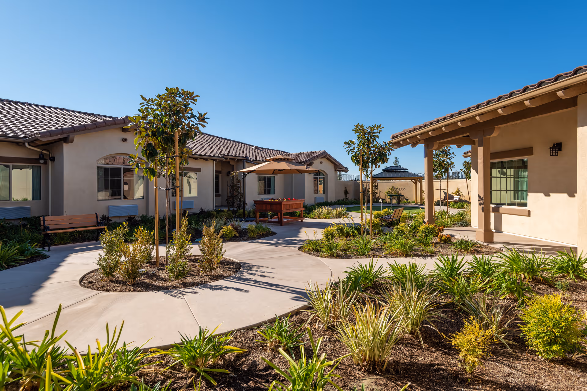 Outdoor courtyard area at ActivCare Laguna Hills featuring a paved walkway winding through landscaped garden beds with small trees and shrubs. There are benches and a shaded table with an umbrella, surrounded by single-story buildings with tiled roofs under a clear blue sky.