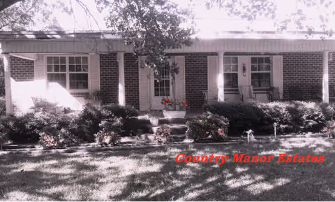 Front exterior view of a single-story brick building with a covered porch supported by white columns. There are two windows and a door on the porch, with some chairs and plants visible. The lawn and shrubs are well-maintained in front of the building.