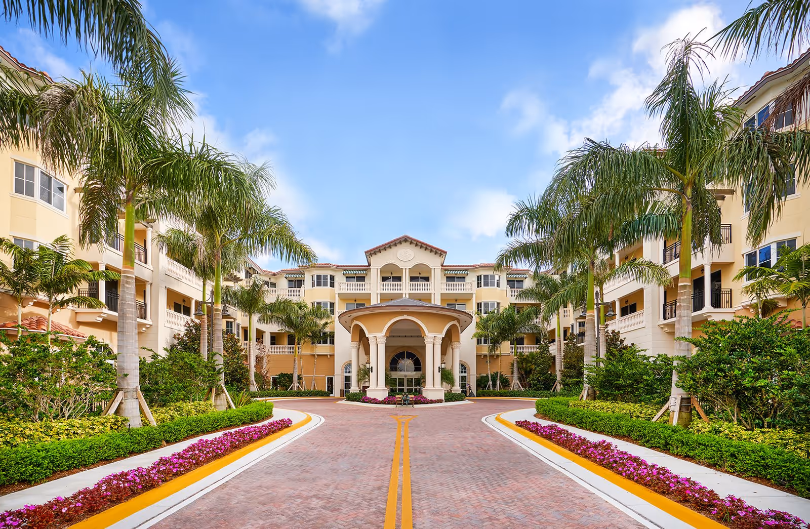 Front exterior view of The Palace at Weston, featuring a grand entrance with a covered portico, surrounded by palm trees, manicured bushes, and colorful flower beds along a brick-paved driveway under a blue sky with some clouds.