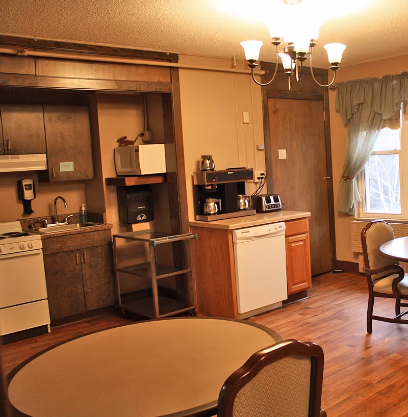 A small kitchen and dining area with wooden floors and beige walls. The kitchen includes a stove, sink, microwave, coffee maker, toaster, and dishwasher. There are two round tables with chairs, a window with blue curtains, and a wooden door. A chandelier with five lights hangs from the ceiling.