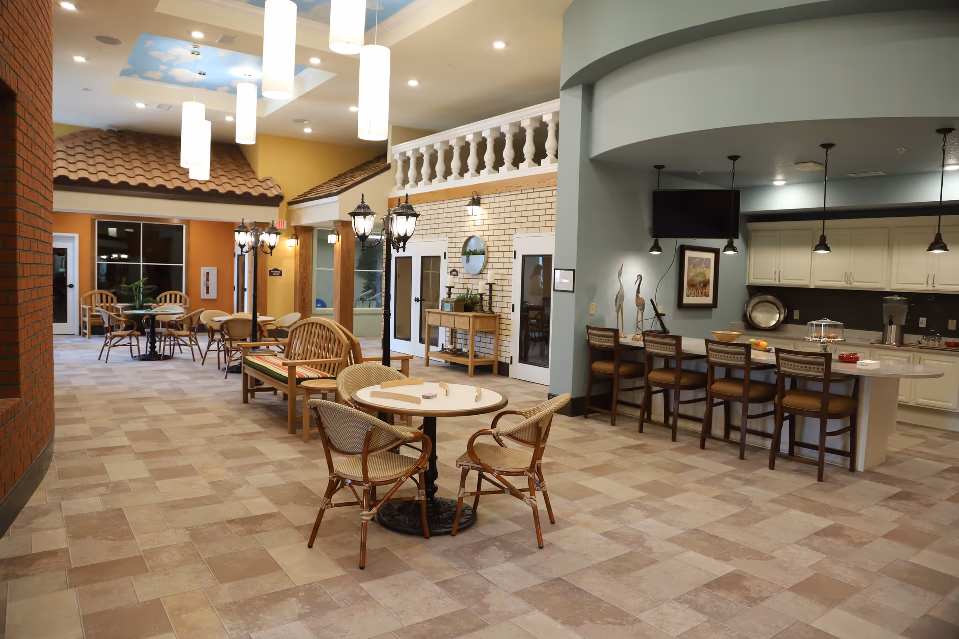 A spacious and well-lit common area in a senior living facility featuring multiple seating arrangements including round tables with chairs, a bench with cushions, and a kitchen counter with bar stools. The area has warm-toned walls, decorative streetlamp-style lights, and a ceiling with a sky mural. The kitchen area includes white cabinets, pendant lights, and a TV mounted on the wall.