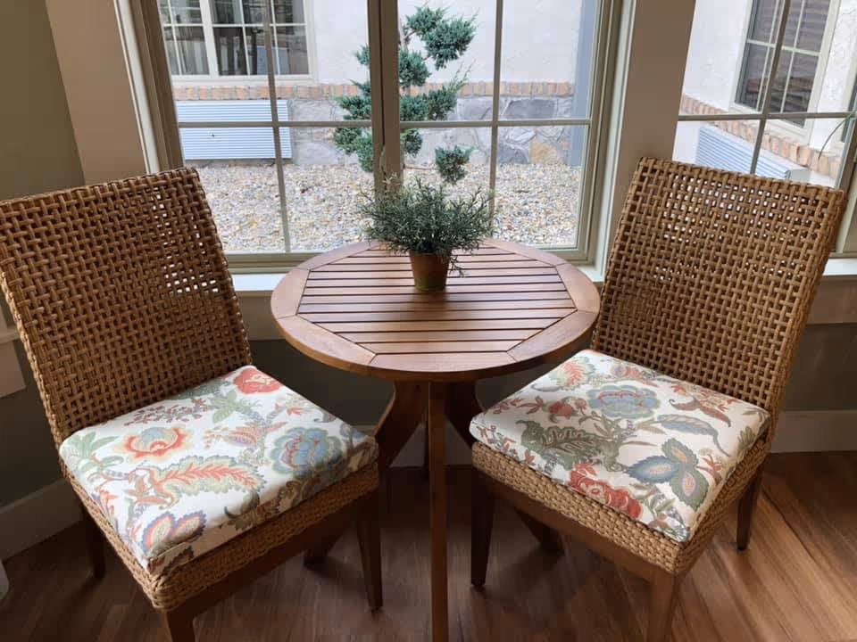 Two wicker chairs with floral cushions flank a small round wooden table holding a potted plant in front of a window.