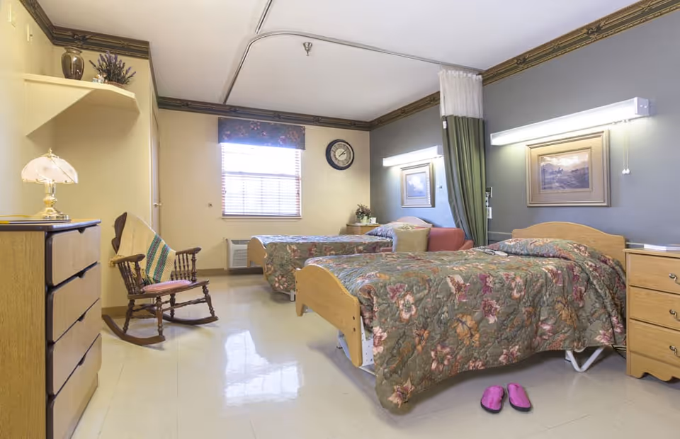 Shared nursing facility bedroom with two floral-covered beds, a rocking chair, dresser and window.