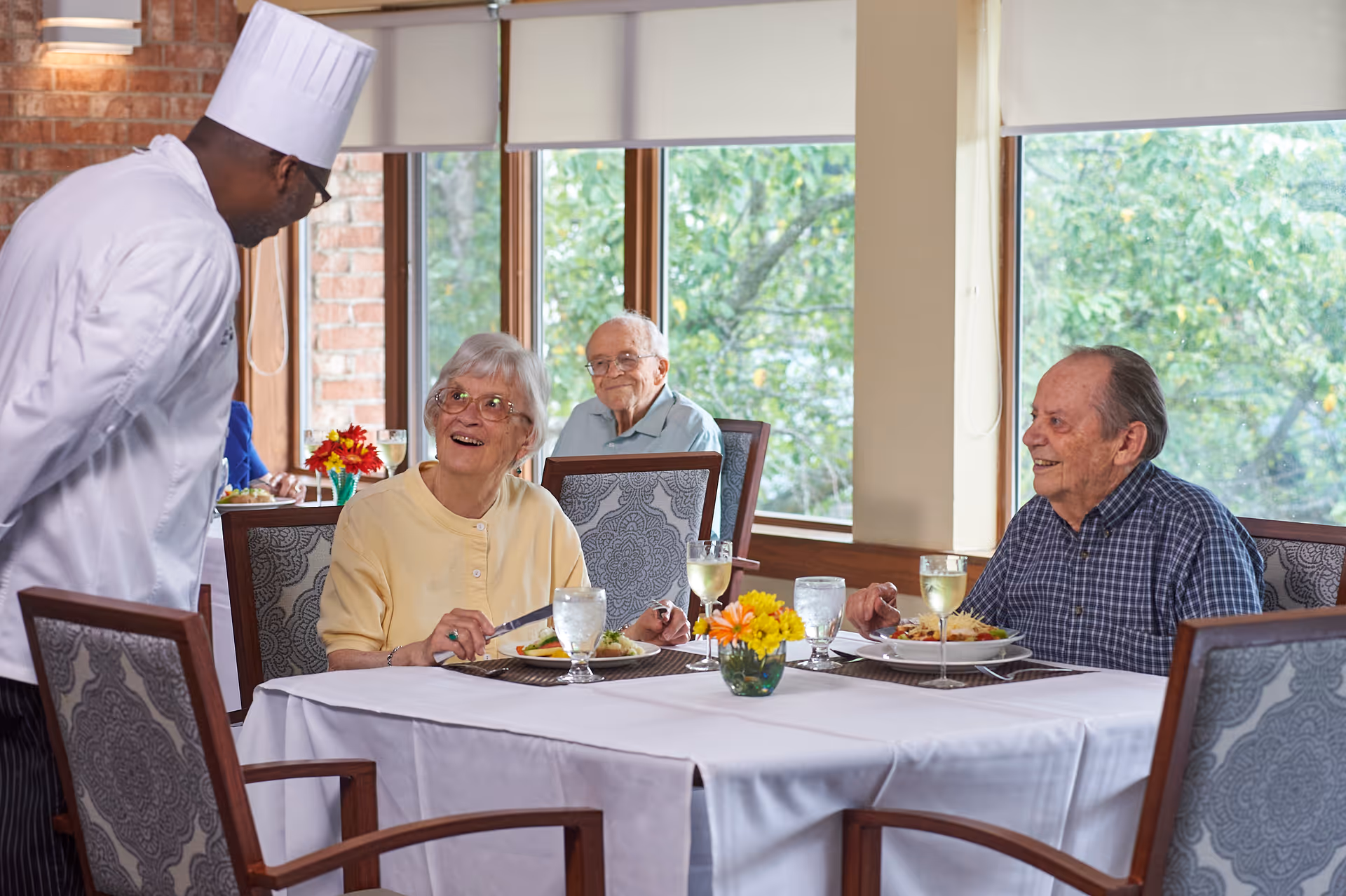 Elderly residents sit at a table in a bright dining room enjoying a meal while a chef talks with them.