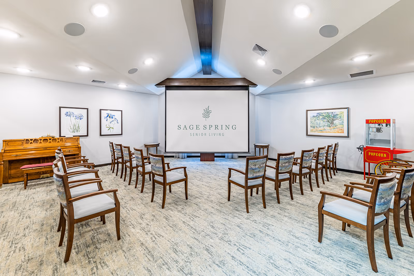 A bright community event room with chairs arranged facing a projection screen that reads 'Sage Spring Senior Living,' a piano on the left and a popcorn machine on the right.