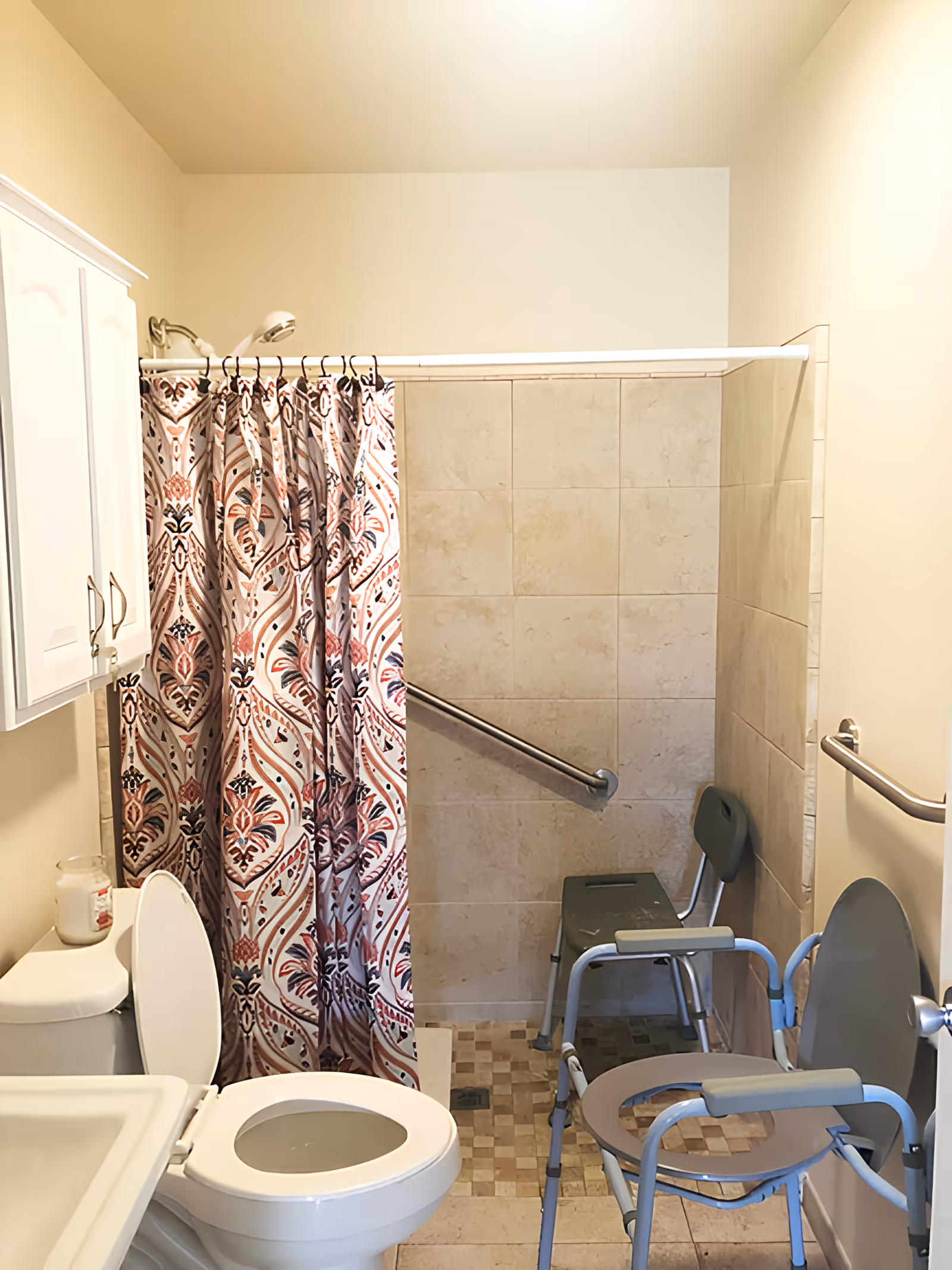 A bathroom with a toilet, a sink, a shower area with a patterned shower curtain, and two shower chairs. The shower area has beige tiles and a grab bar for support. There is also a white cabinet mounted on the wall above the toilet.