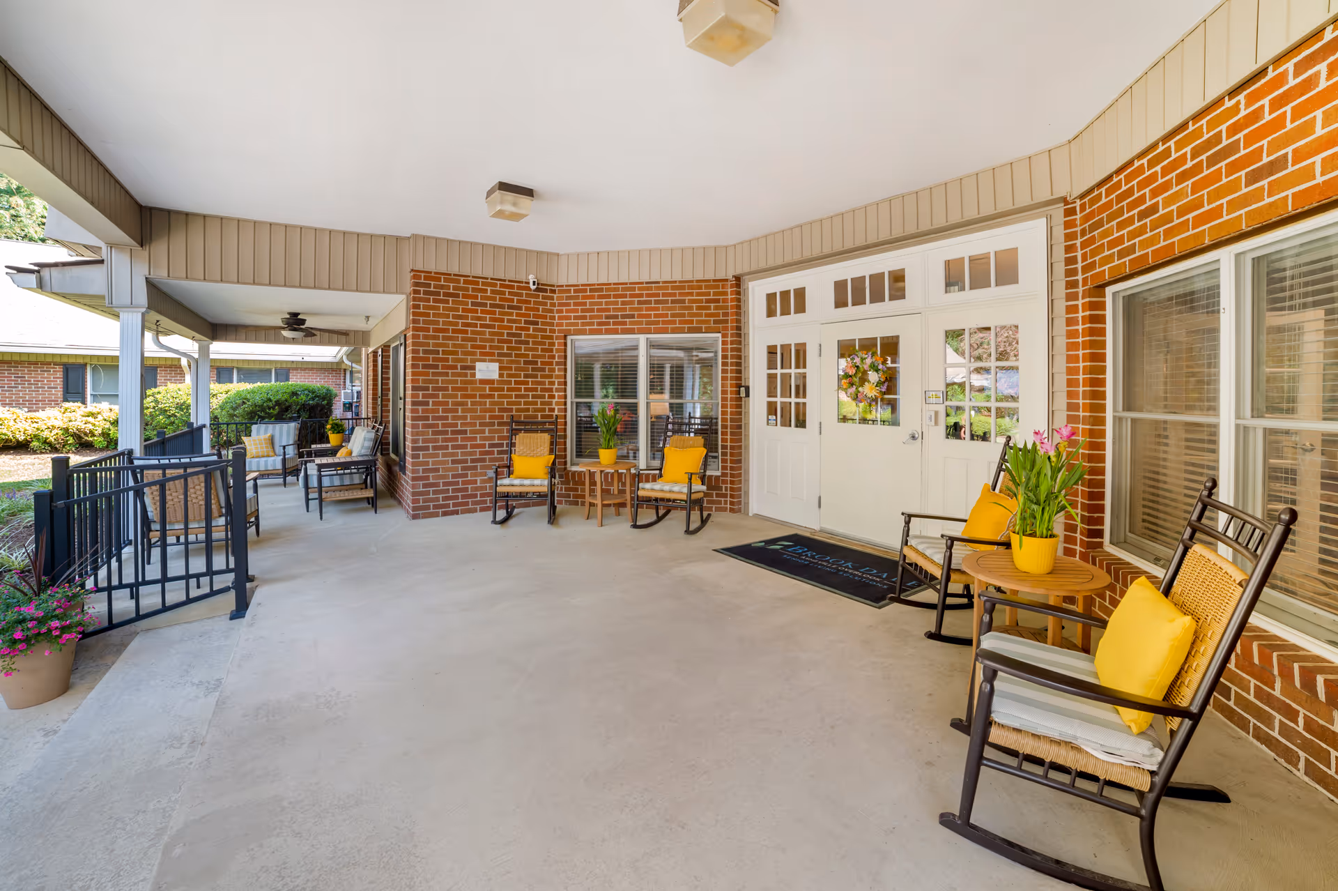 Covered entrance porch with rocking chairs, yellow cushions, potted plants, and white double doors on a brick building.
