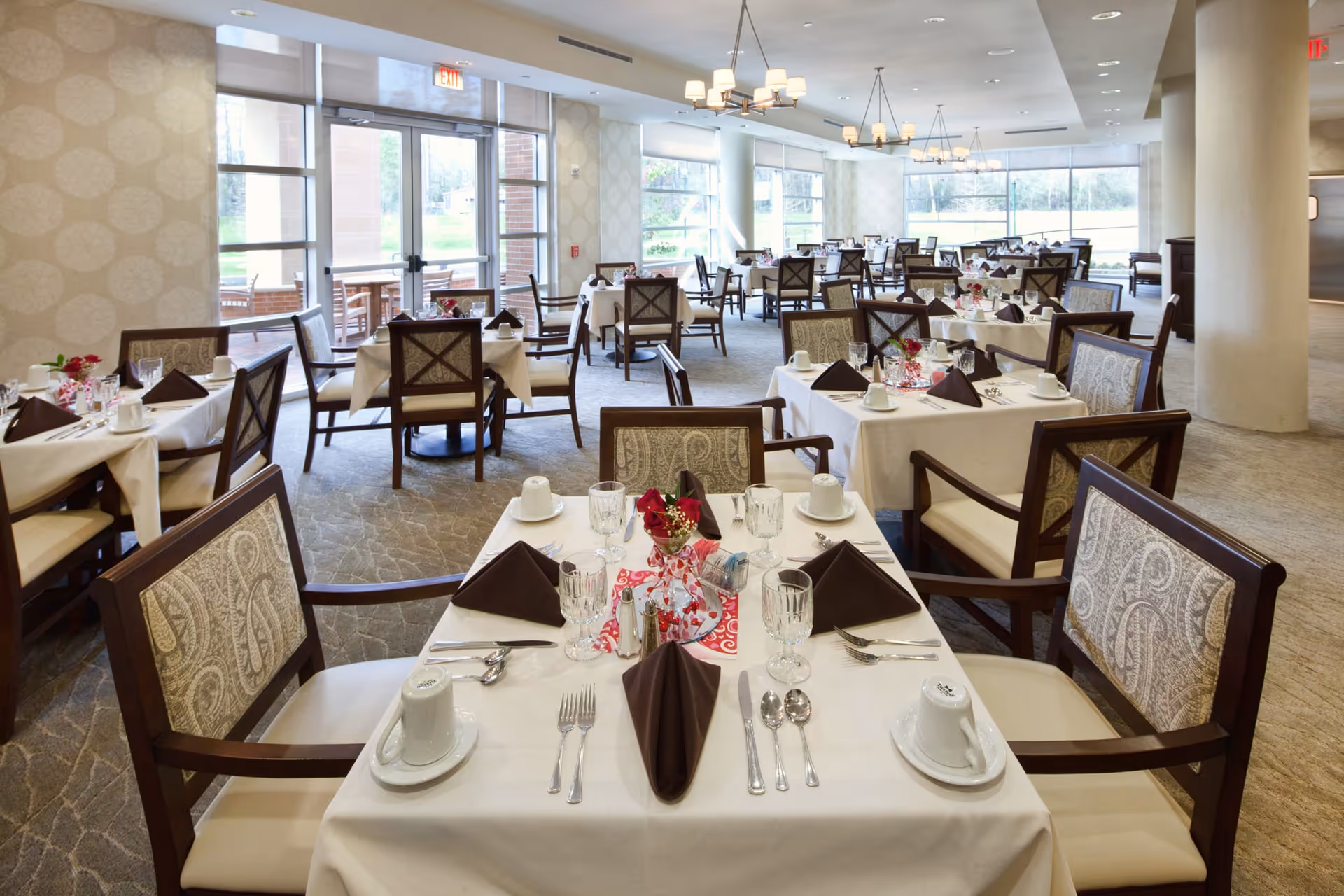 A spacious dining room with multiple tables covered in white tablecloths, each set with brown napkins, glassware, silverware, and small floral centerpieces. The room has large windows letting in natural light and elegant chandeliers hanging from the ceiling.