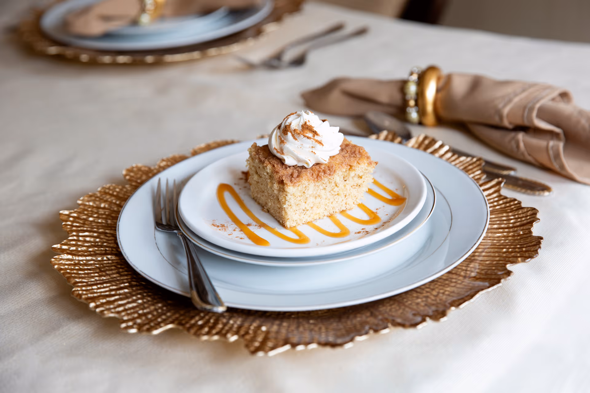 A close-up of a dessert plate with a square piece of cake topped with whipped cream and a sprinkle of cinnamon, placed on a white plate with caramel sauce drizzled around it. The plate is set on a gold decorative charger with a fork resting on the side. In the background, there is a beige napkin with a gold napkin ring and additional table settings.