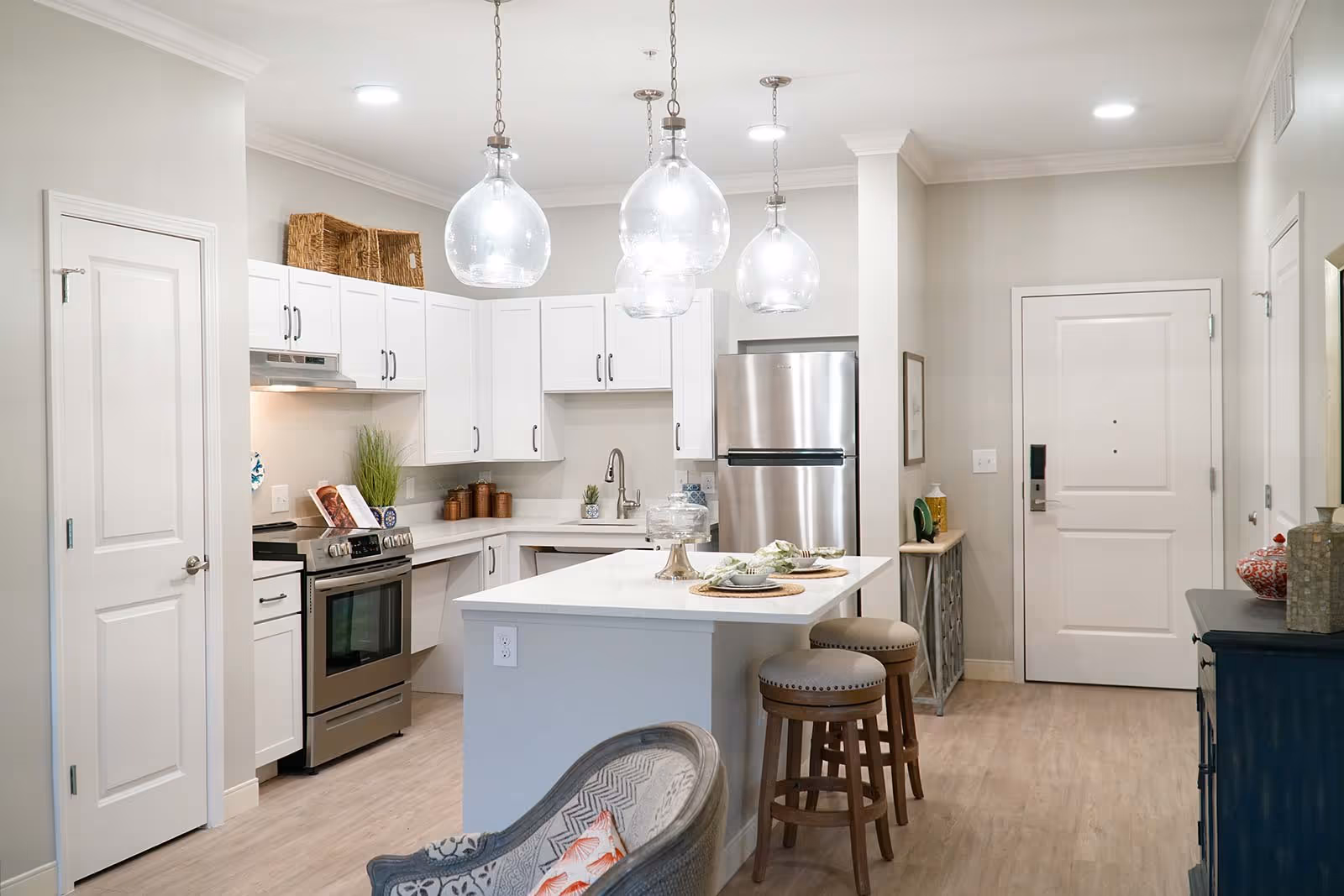 Modern kitchen with white cabinets, stainless steel appliances including a refrigerator and stove, a white island with two wooden stools, and four glass pendant lights hanging from the ceiling. The kitchen is adjacent to an entry door and a small console table with decorative items.