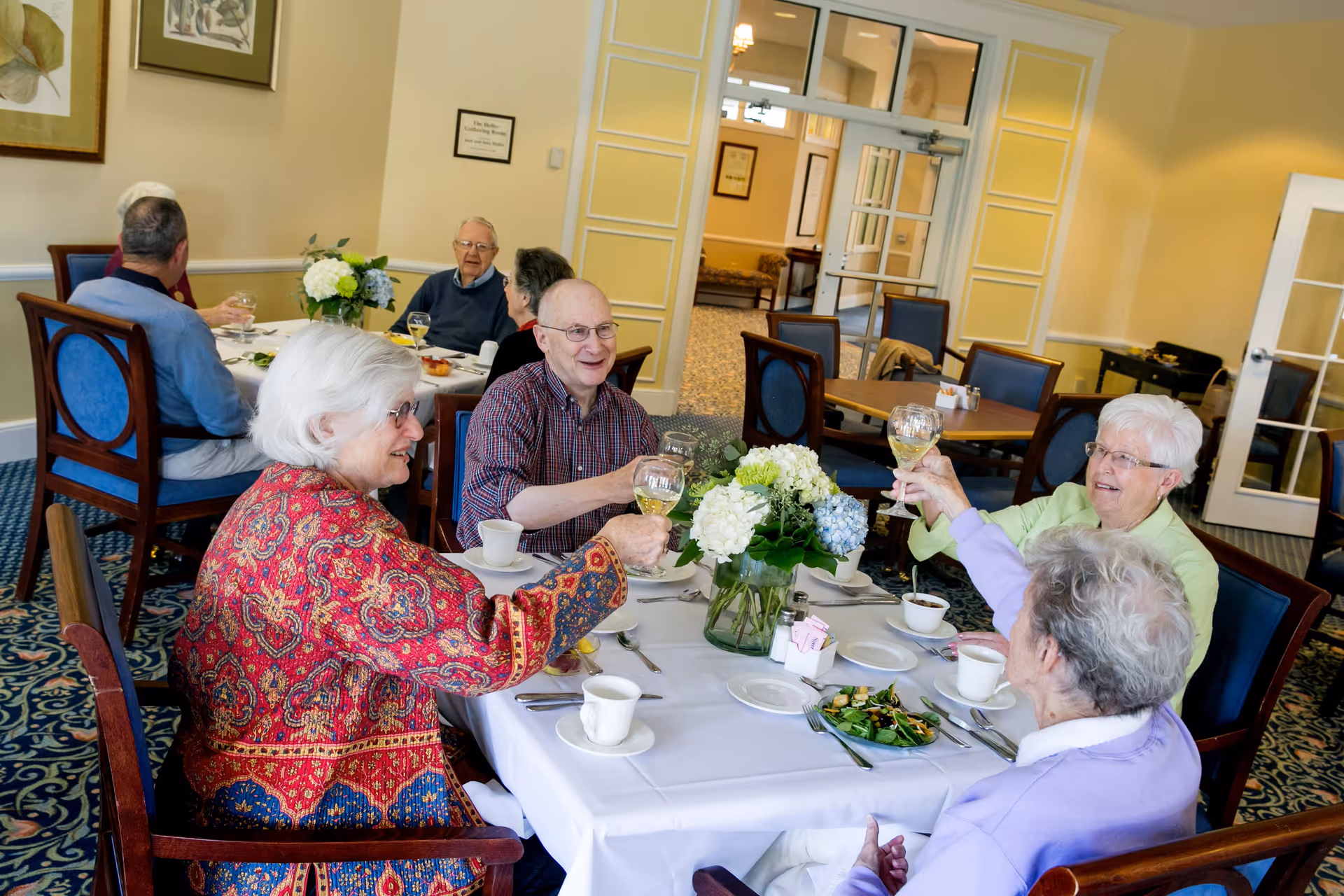 A group of elderly people sitting around a dining table in a senior living community, raising glasses in a toast. The table is set with white tablecloth, cups, plates, and a vase of white and blue hydrangeas. The room has yellow walls, blue carpet, and wooden chairs with blue cushions.