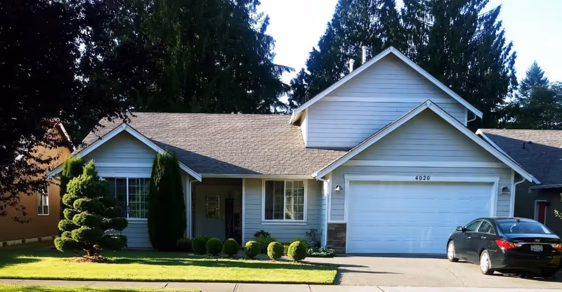 Front exterior view of a single-story house with light gray siding, a two-car garage, a neatly trimmed lawn, and a black car parked in the driveway. The house number 4020 is visible above the garage door, and tall trees are in the background.