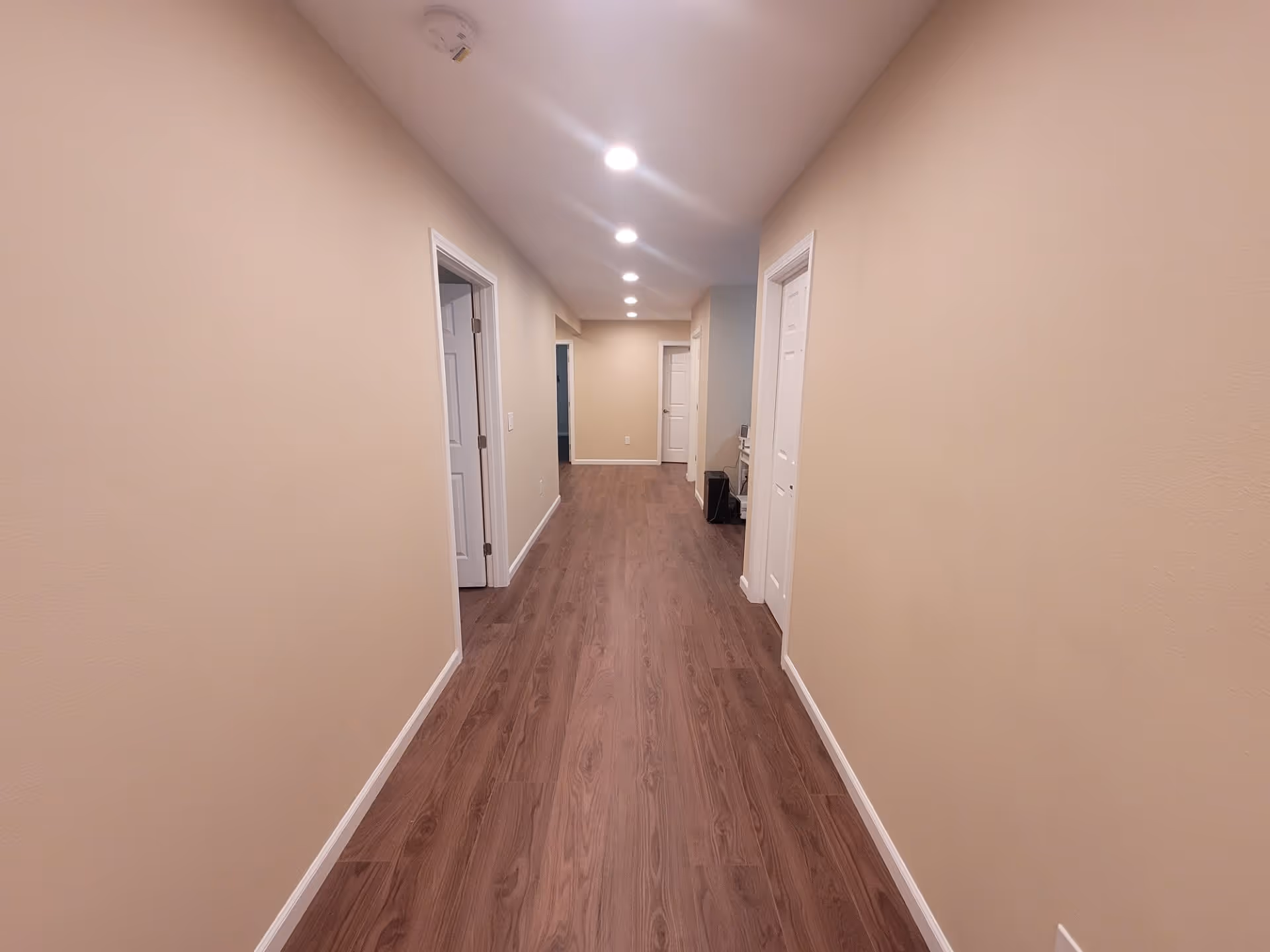 A long, narrow hallway with wooden flooring and beige walls. The hallway has several white doors on both sides and recessed ceiling lights evenly spaced along the ceiling. A smoke detector is visible on the ceiling near the foreground.