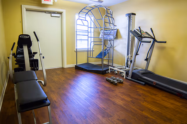 Small fitness room with wooden flooring and yellow walls containing exercise equipment including a treadmill, a multi-use gym machine, a bench press, and a set of dumbbells near a window and a closed door.