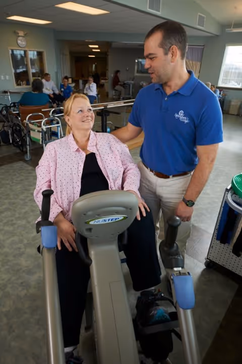 A woman seated on a NuStep exercise machine in a rehabilitation or therapy room, smiling and looking up at a man standing beside her who is wearing a blue polo shirt. In the background, other people are engaged in activities and there are wheelchairs and exercise equipment visible.