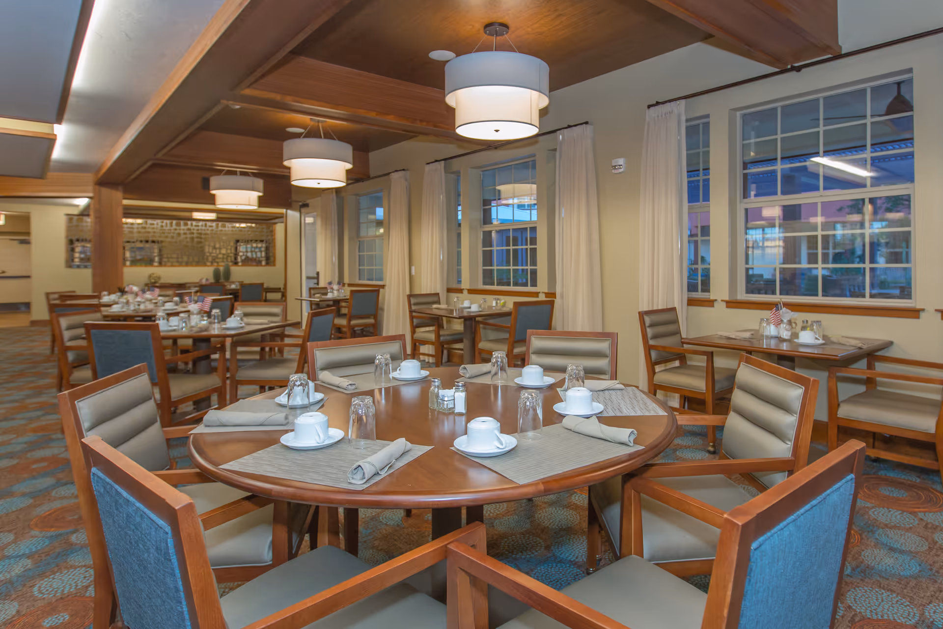 Dining room with round wooden tables set with cups and napkins, upholstered chairs, pendant lights, and large windows.