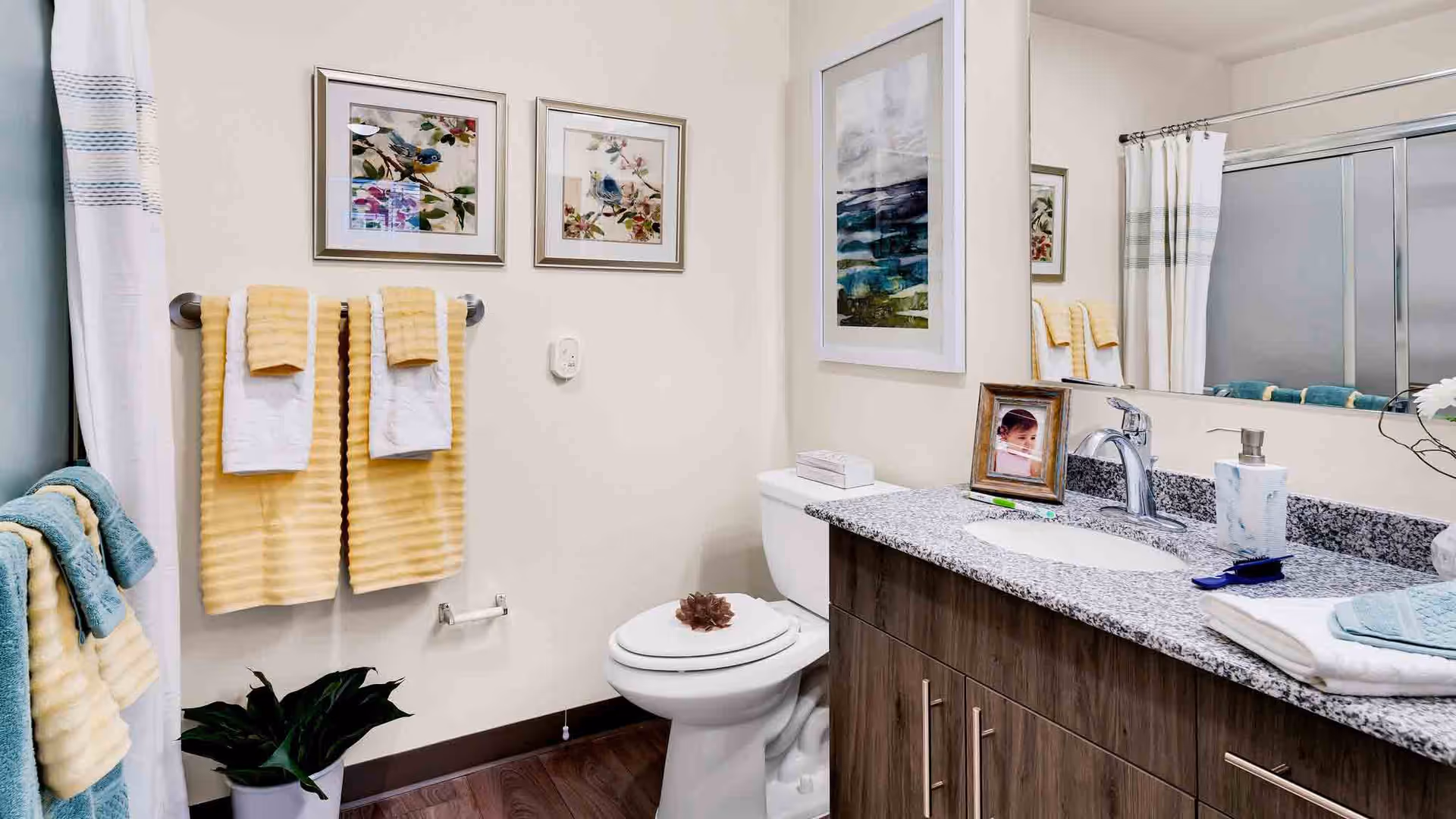 A clean and well-decorated bathroom featuring a toilet, a granite countertop with a sink, a large mirror, and a shower with a sliding glass door. The bathroom has yellow and white towels hanging on racks, framed floral artwork on the walls, and a small plant near the toilet.