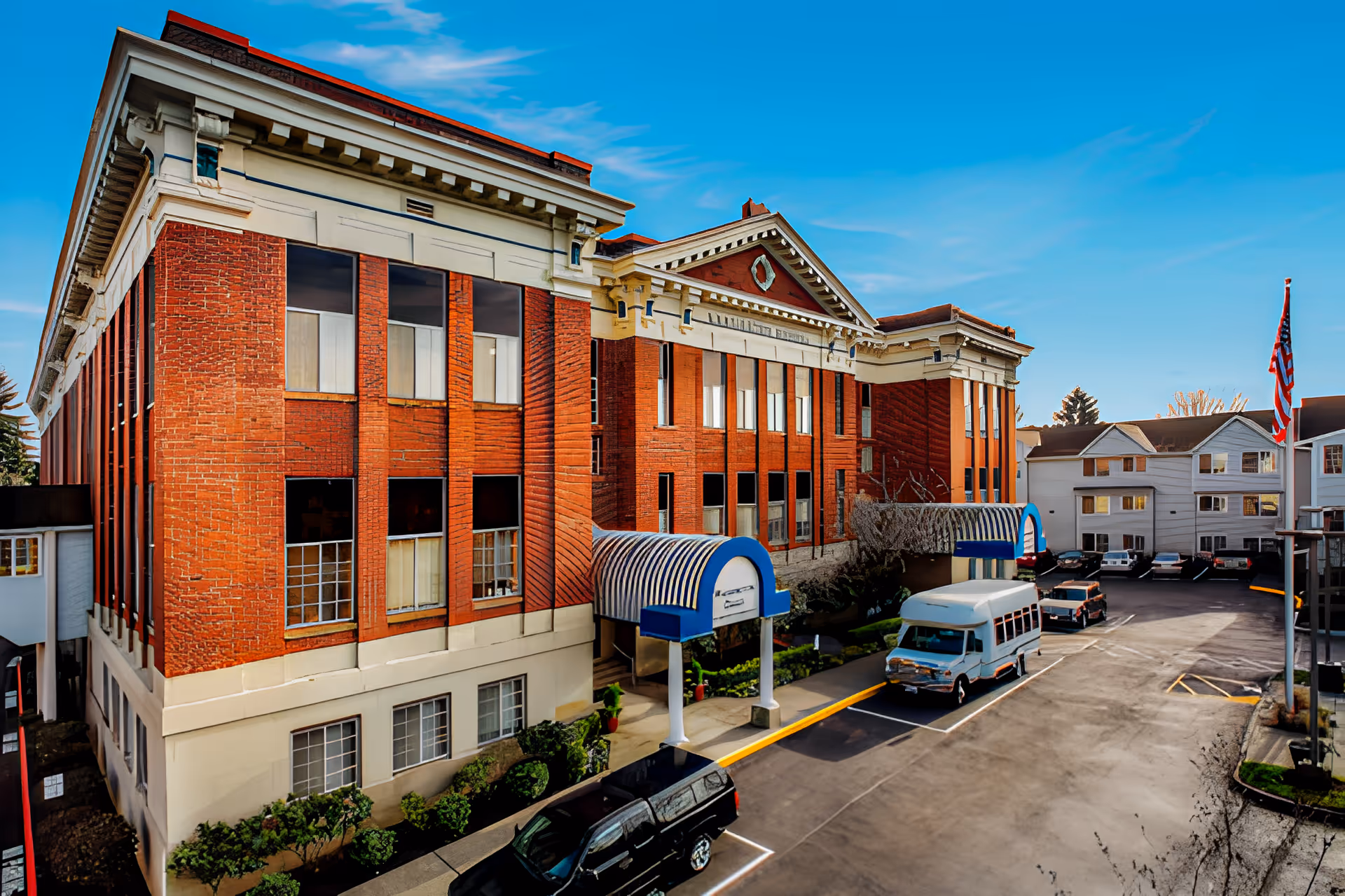 Exterior view of a large red brick building with white trim and blue awnings over entrances. Several vehicles, including a white shuttle bus and black SUV, are parked in the lot. An American flag is visible on a flagpole to the right, and the sky is clear and blue.