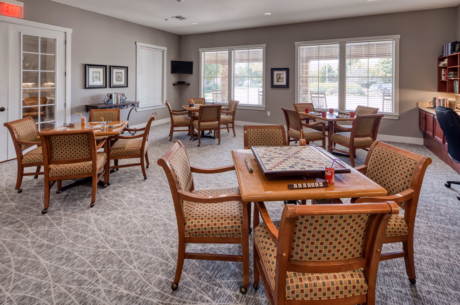 A bright and spacious common room with multiple wooden tables and cushioned chairs arranged for group activities. One table in the foreground has a Scrabble board game set up, along with a can of Coca-Cola and a juice box. Large windows with blinds allow natural light to fill the room. The walls are painted light gray and decorated with framed pictures. A small TV is mounted in the corner, and a desk with books and a chair is visible on the right side.