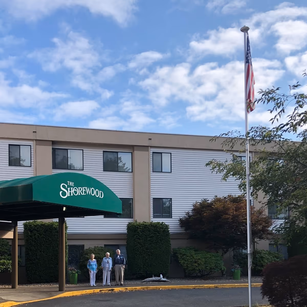 Exterior view of The Shorewood senior living facility with a green awning over the entrance. Three elderly people stand near the entrance. There is an American flag on a flagpole to the right, and bushes and trees surround the building under a partly cloudy sky.