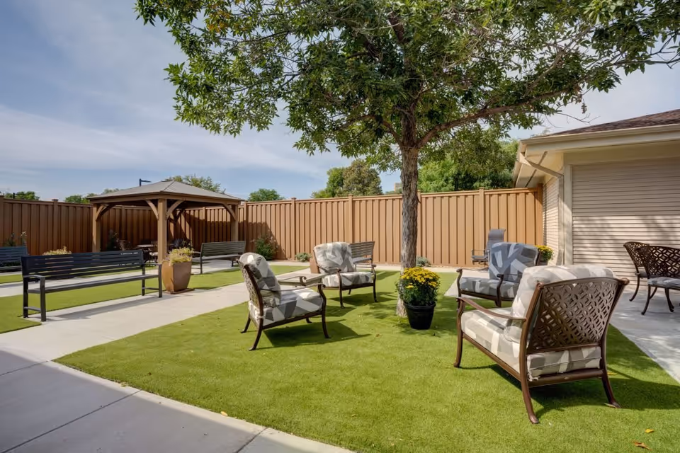 Outdoor seating area at The Courtyards at Mountain View featuring cushioned chairs arranged around a tree with potted plants, benches, a gazebo, and a fenced yard under a clear sky.