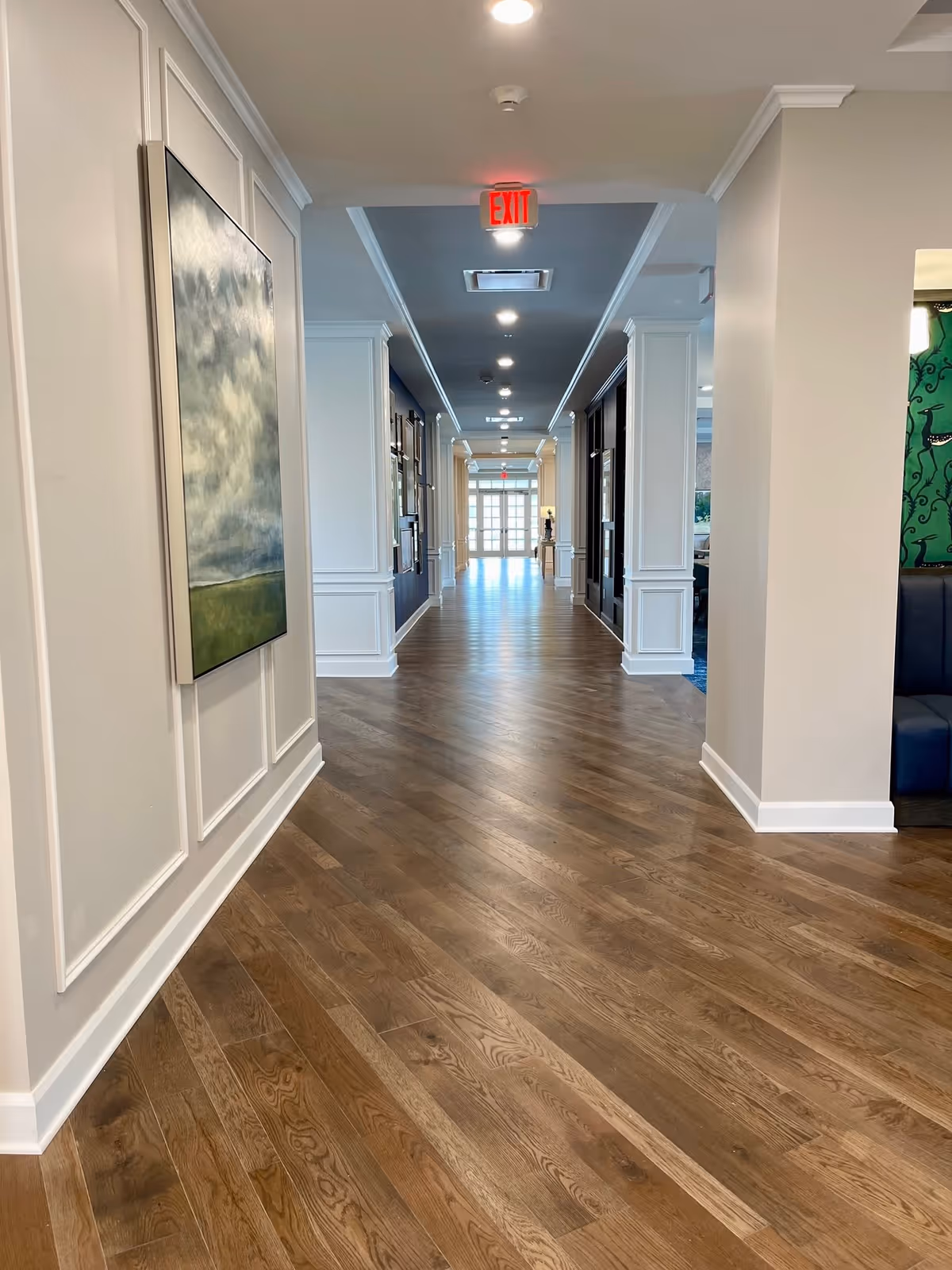 Well-lit interior hallway with wood floors, decorative wall panels and artwork, columns lining the sides, and an illuminated EXIT sign at the ceiling.