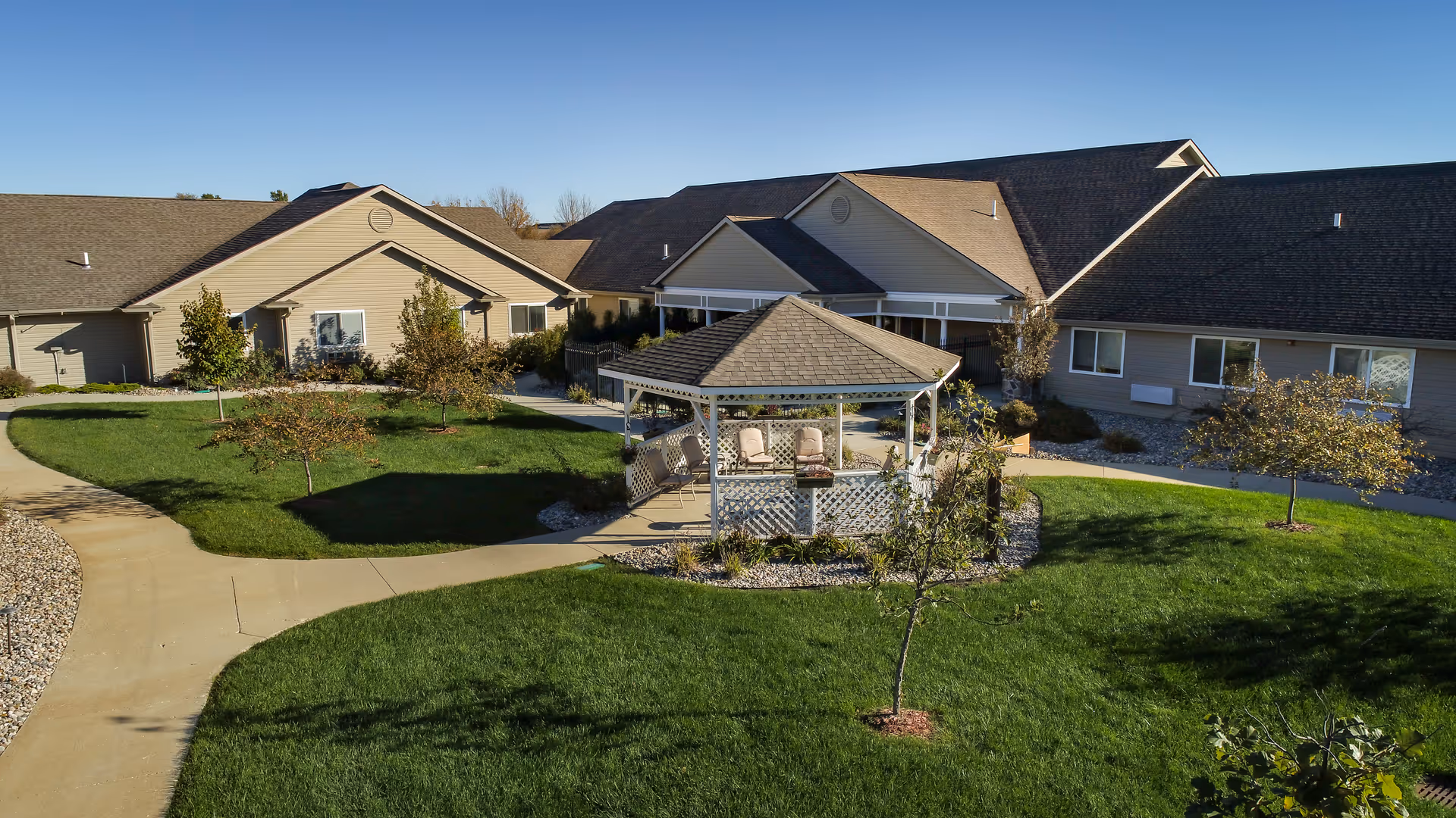Landscaped courtyard with a central gazebo, walkways, small trees, and surrounding single-story assisted living buildings under a clear sky.
