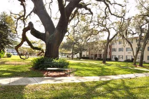 Exterior view of a senior living facility with lawn, trees, and a bench.