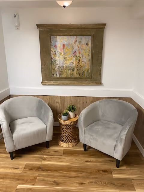 Two gray upholstered armchairs positioned on either side of a small round wicker table with three small potted plants on it. Behind the chairs is a wooden wainscoting with white walls above, and a framed painting of flowers hanging on the wall.