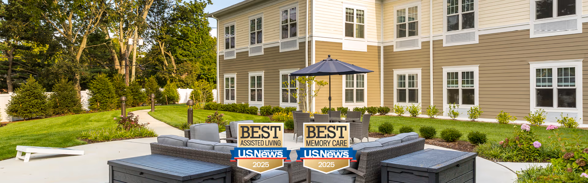 Outdoor seating area at Orchard Estate of Woodbury featuring modern wicker furniture with cushions arranged around a table with an umbrella. Two signs display awards for Best Assisted Living and Best Memory Care by U.S. News for 2025. The area is surrounded by well-maintained landscaping and a beige multi-story building with white-trimmed windows in the background.