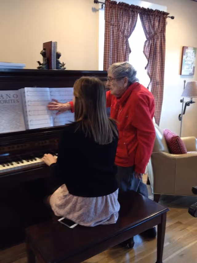 An elderly woman in a red jacket is standing next to a younger woman seated at a piano. The elderly woman is pointing at sheet music on the piano while the younger woman plays. The room has a window with checkered curtains, a beige armchair with a red pillow, and a floor lamp with a checkered lampshade.