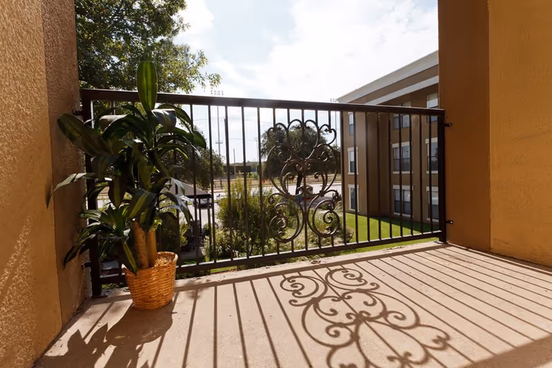 A small balcony with a decorative black metal railing casting intricate shadows on the floor. A potted green plant is placed in the corner of the balcony. In the background, there is a view of a grassy area, trees, and a multi-story building under a partly cloudy sky.