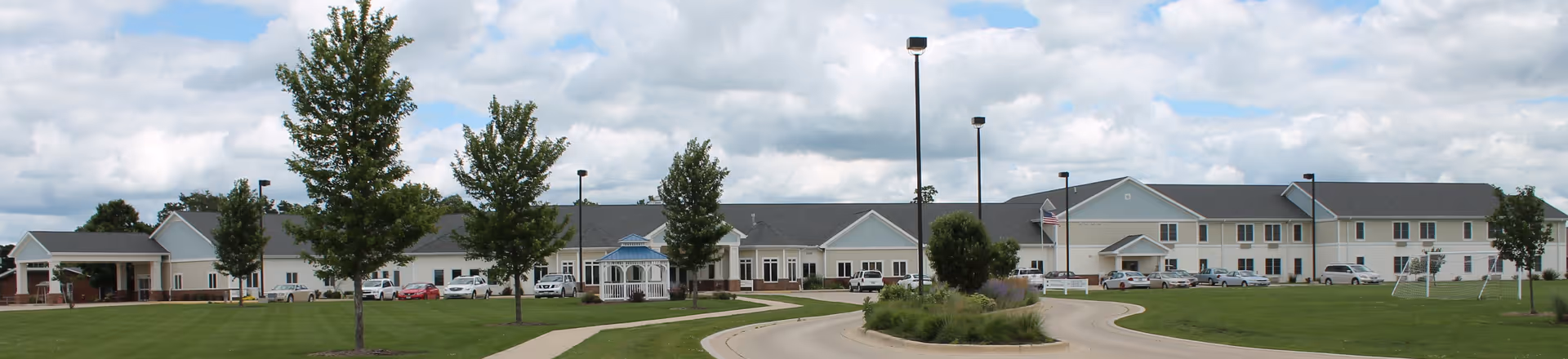 Wide exterior view of a large, single-story and two-story senior living facility with a well-maintained lawn, several trees, a gazebo, and a circular driveway under a partly cloudy sky.