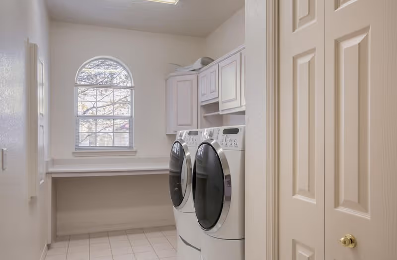 Laundry room with a front-loading washing machine and dryer side by side, white cabinetry above, a countertop along the wall, and a window with an arched top letting in natural light.