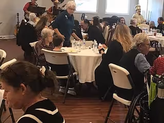 A group of elderly people seated around tables in a dining area, engaging in conversation and being served by a staff member. The room has wooden flooring and large windows letting in natural light.