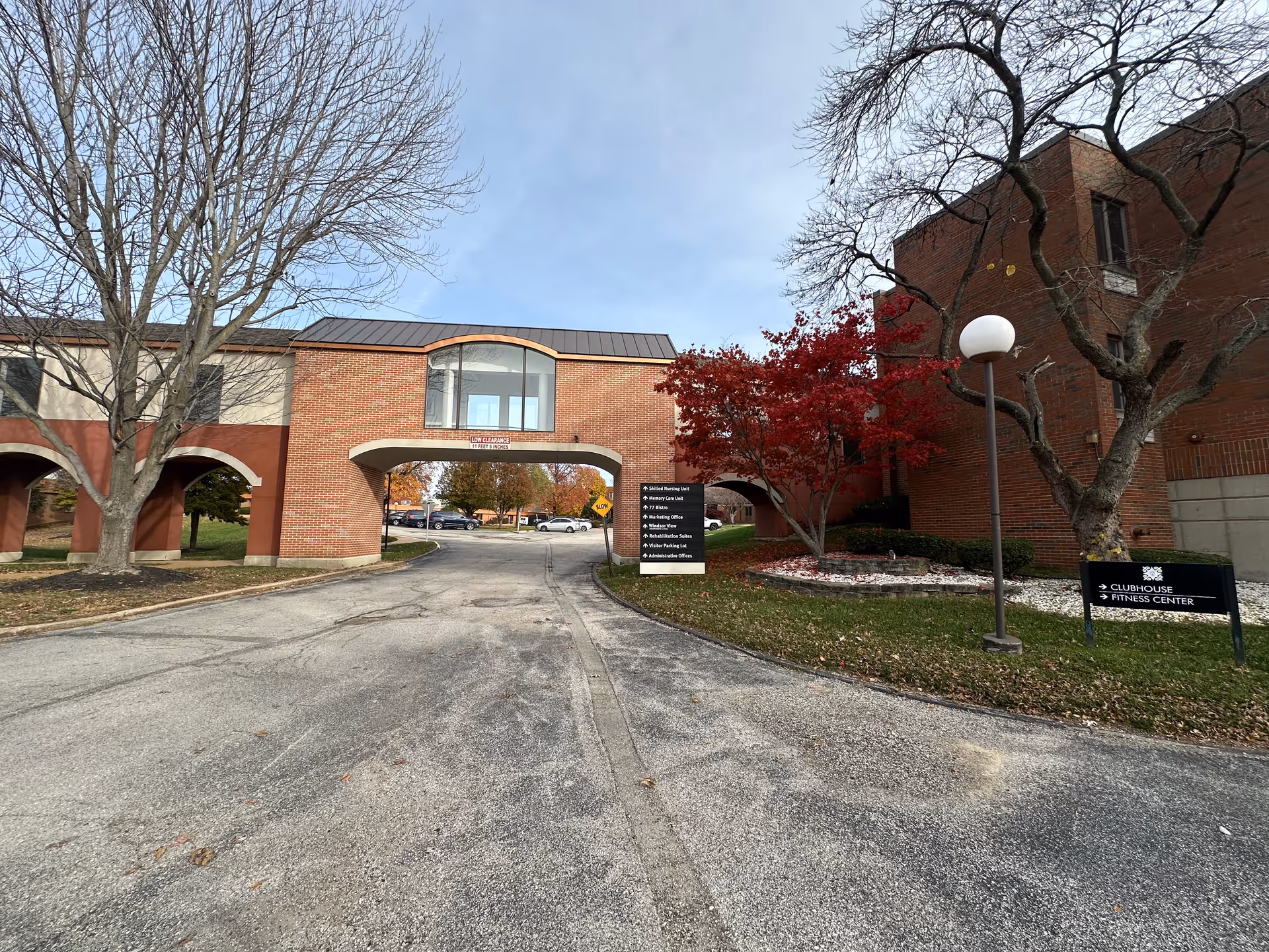 Exterior view of a senior living facility with a brick archway over a driveway. There are leafless trees and a tree with red leaves near the buildings. Signs indicate directions to various units and amenities including Skilled Nursing Unit, Memory Care Unit, Marketing Office, Windsor View, Rehabilitation Suites, Visitor Parking Lot, Administrative Offices, Clubhouse, and Fitness Center. The sky is clear with some light clouds.