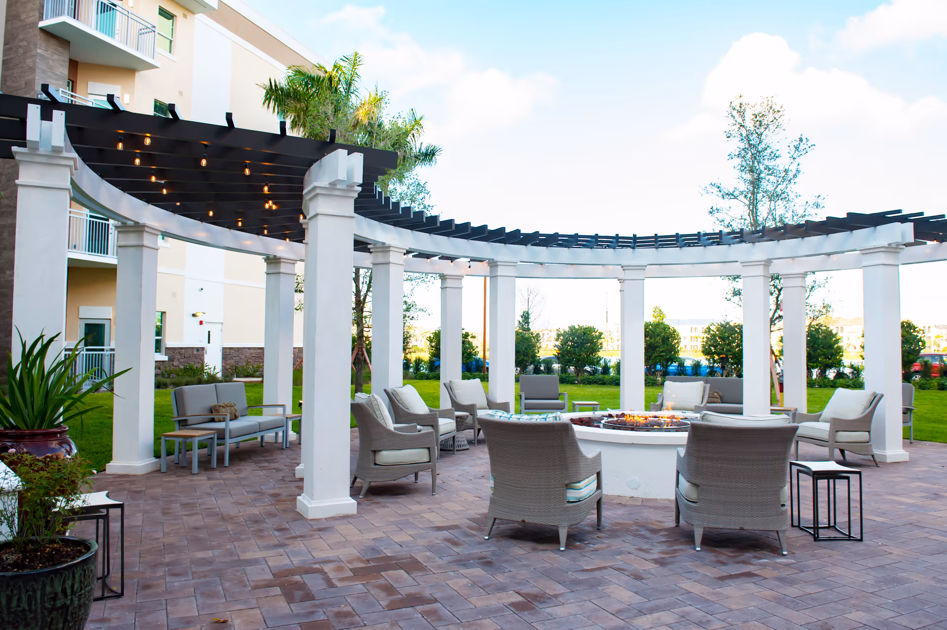 Outdoor courtyard with a white pergola and cushioned chairs arranged around a central fire pit on a brick patio.
