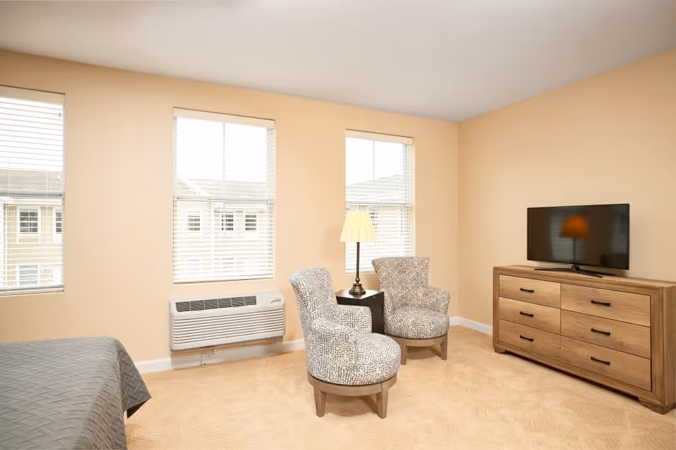 A bright room with beige walls and carpet featuring two patterned armchairs around a small table with a lamp, a wooden dresser with a flat-screen TV on top, three windows with white blinds, and a portion of a bed with a gray quilt visible on the left side.