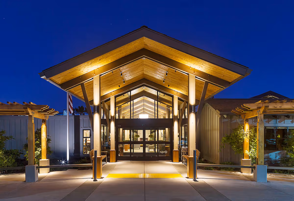 Entrance of The Redwoods retirement facility at night, featuring a well-lit wooden canopy with glass doors, benches on either side, and landscaped surroundings under a clear dark blue sky.
