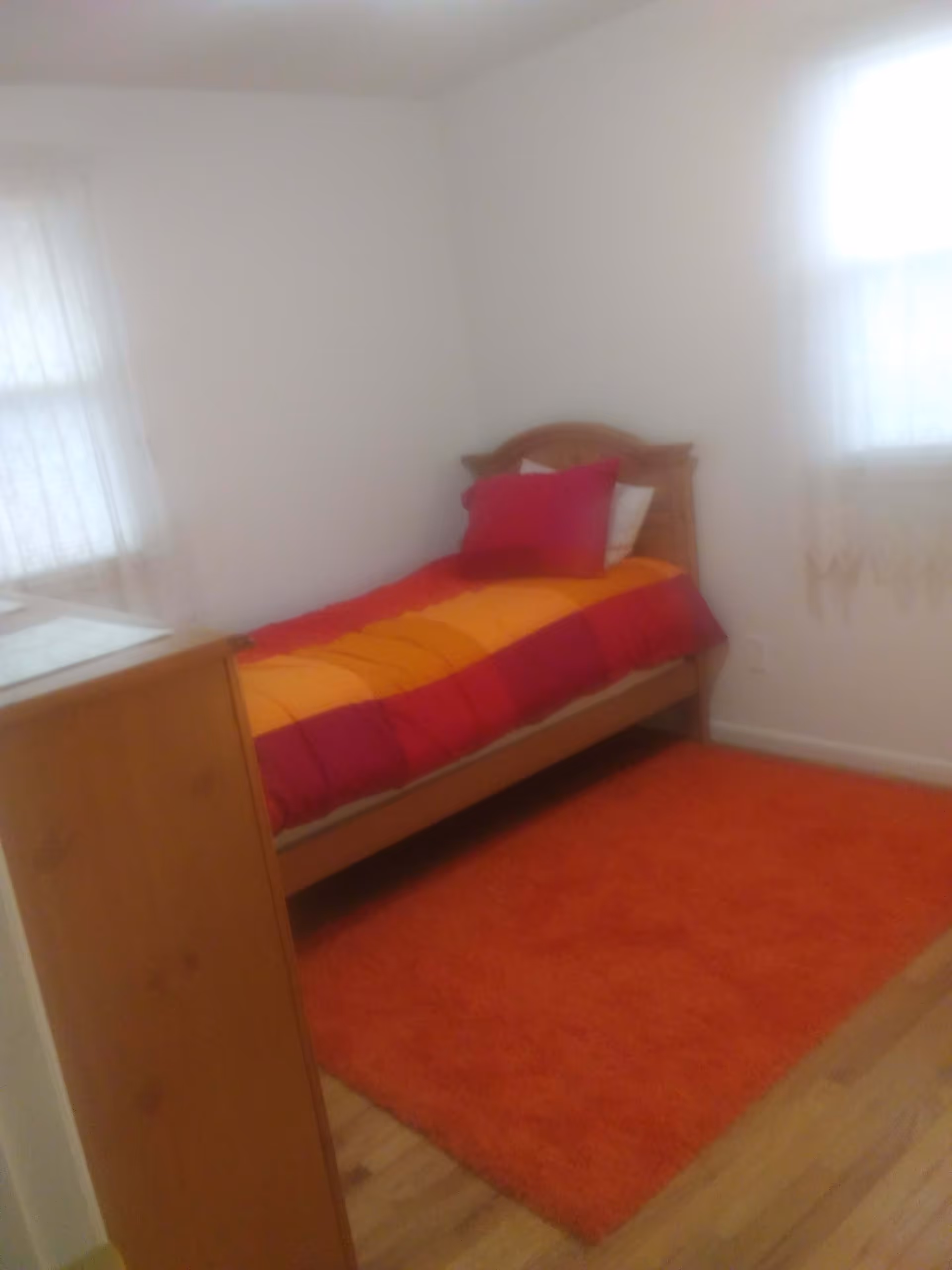 A small bedroom with a single wooden bed against the wall, covered with a red and orange striped blanket and a red pillow. There is a bright orange rug on the wooden floor and two windows with sheer white curtains letting in natural light.