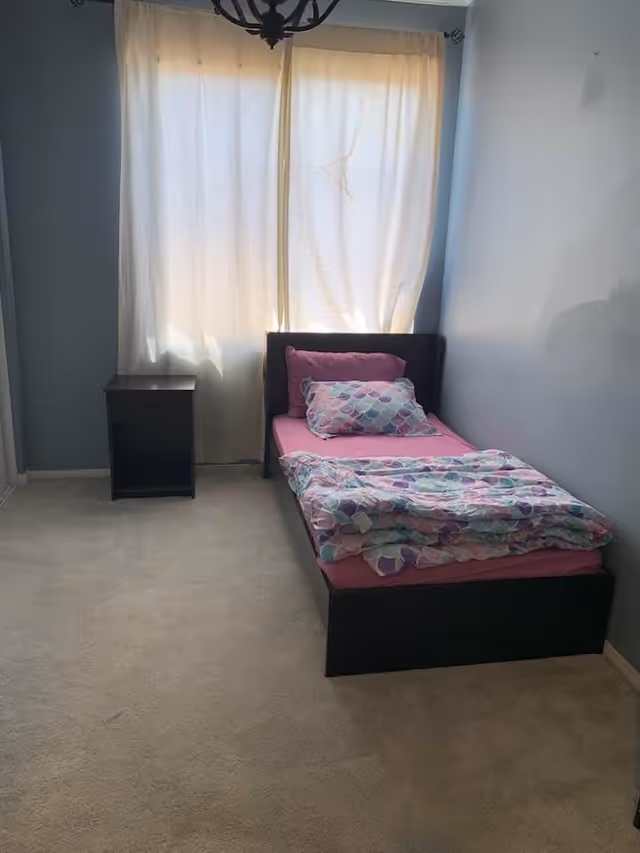 Small bedroom with a single bed dressed in pink sheets and a patterned comforter, a nightstand, and a curtained window.