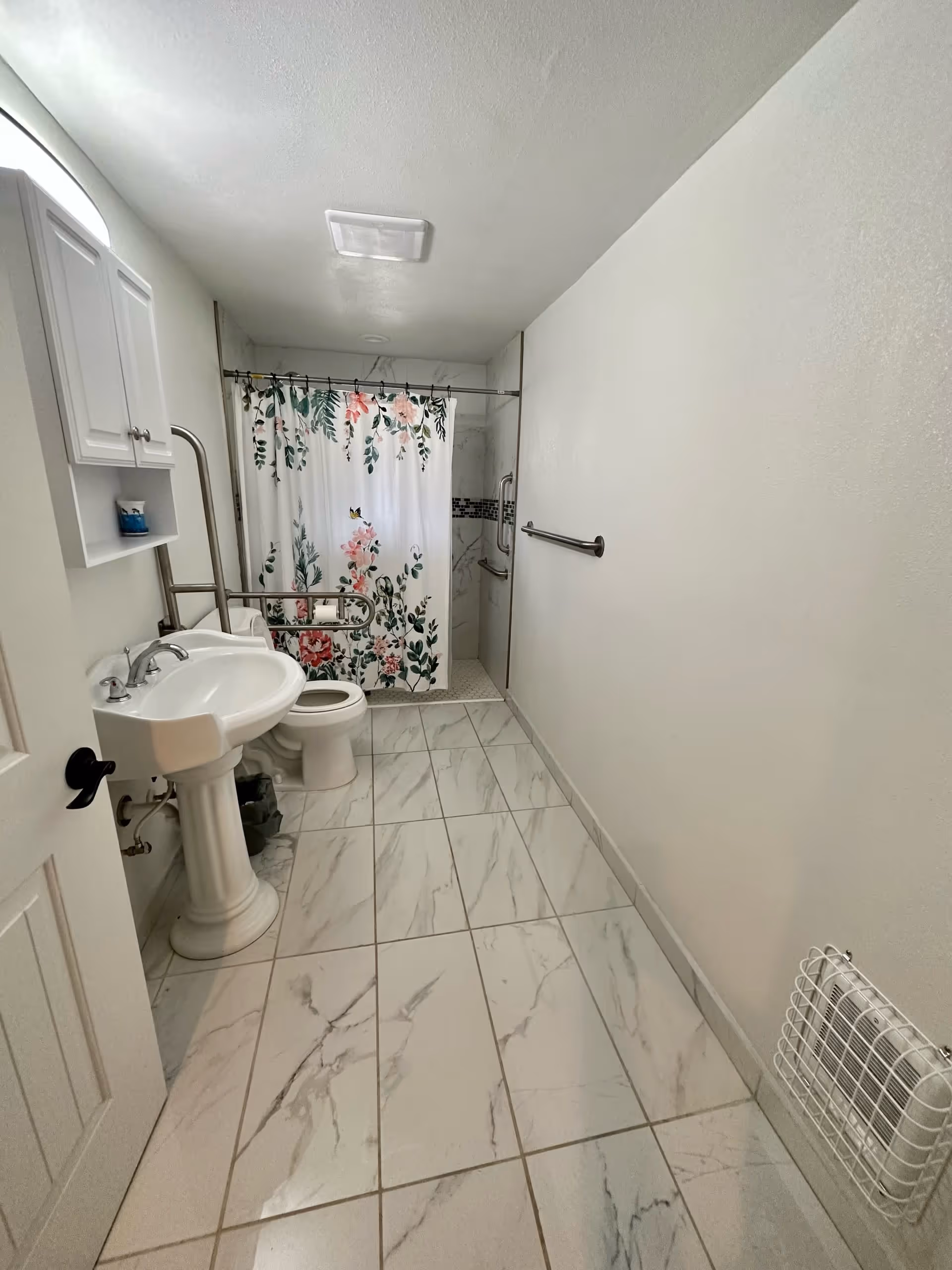 A clean bathroom with white marble-patterned floor tiles, a white pedestal sink, a toilet with safety grab bars, a floral shower curtain, and a wall-mounted heater with a protective cage.