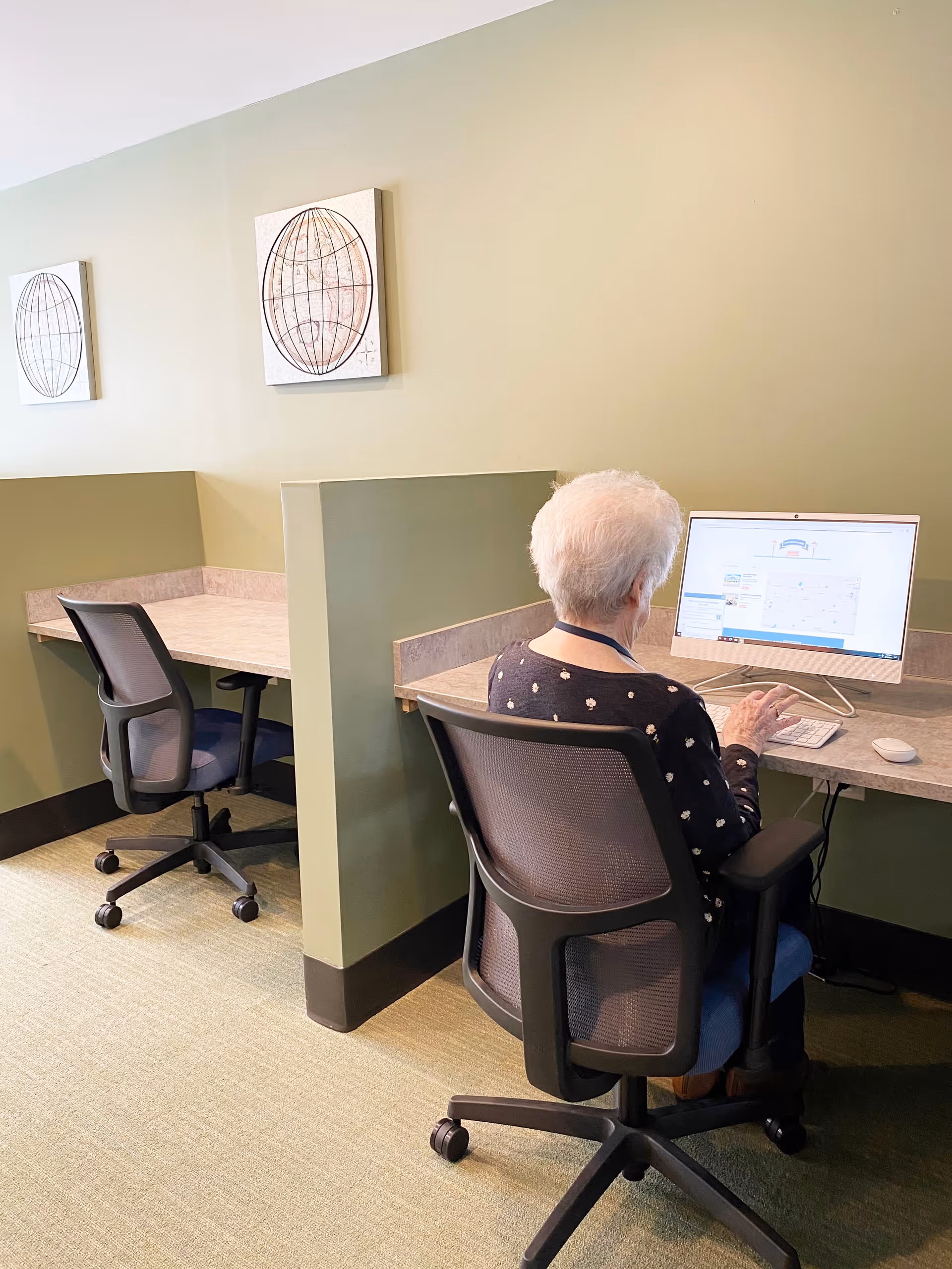 An elderly woman with white hair sits in a black mesh office chair at a computer workstation with a beige countertop and green walls. The workstation is divided by a partial wall, and there is an empty chair and desk space next to her. Two framed globe artworks hang on the wall above the desks.
