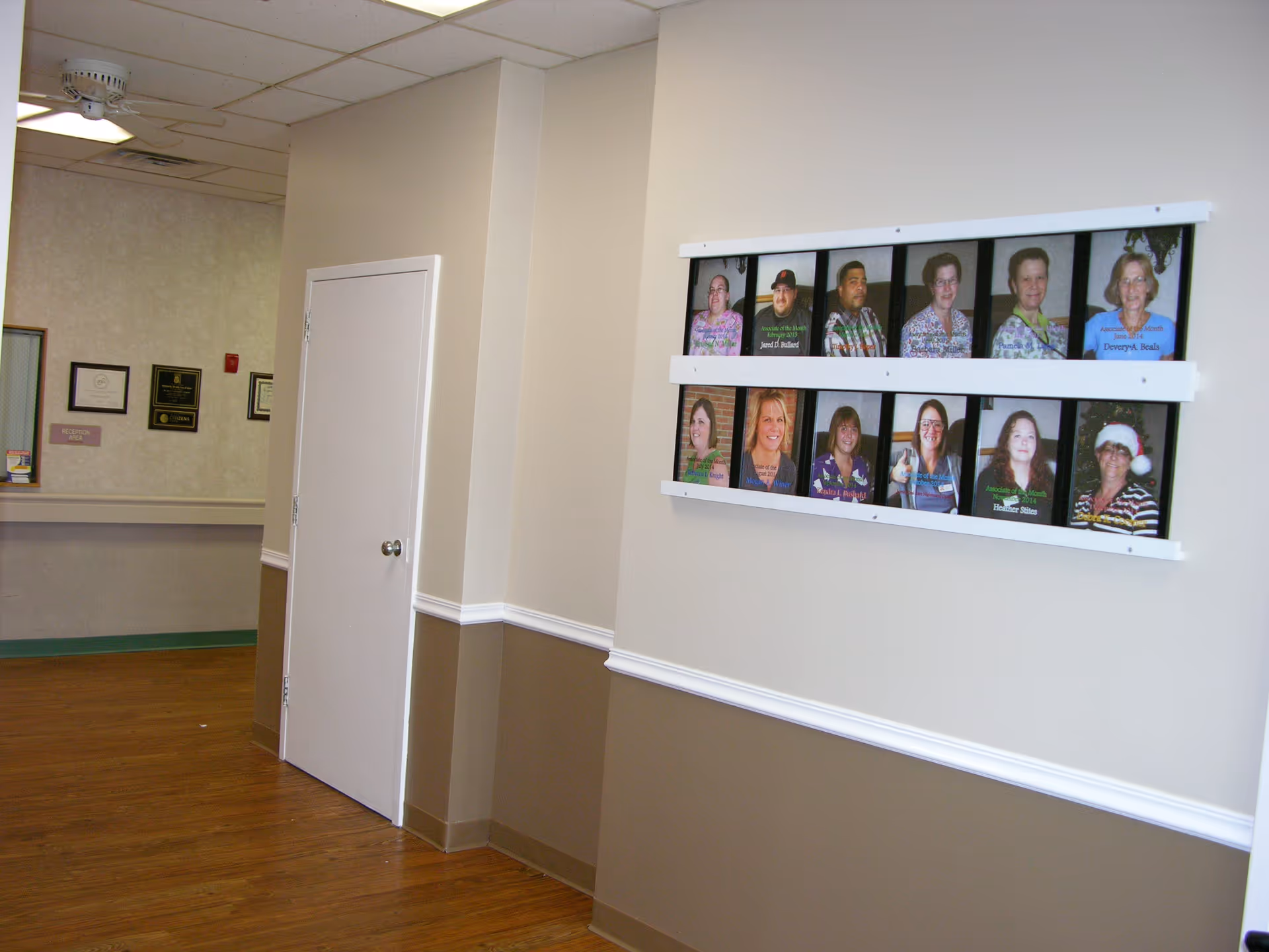 Interior hallway of a senior living facility with beige and brown walls, a white door, and a bulletin board displaying framed photos of staff members on the right wall. The floor is wood, and there are certificates and a reception window visible in the background.